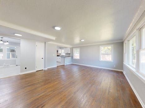 Sunlit empty living room with wooden flooring and large windows.